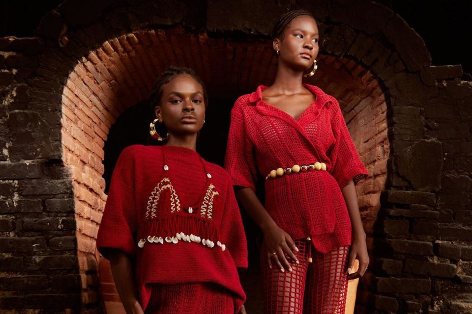 Campanha de divulgação da coleção de roupas da marca Ateliê Mão de Mãe. As modelos, duas mulheres jovens e negras de cabelo trançado, estão em um corredor de tijolos de barro. Vestem um conjunto de crochê vermelho e e acessórios feitos de barro.
