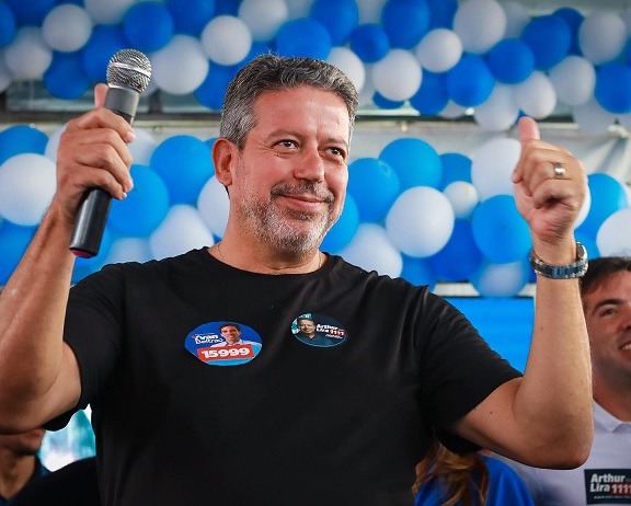Arthur Lira, presidente da Câmara dos Deputados, sorri em cima de palanque durante campanha. Ele segura microfone na mão esquerda e atras é possívle ver balões azuis e brancos - Metrópoles