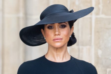 foto colorida de meghan markle usando roupa preta e um chapéu preto