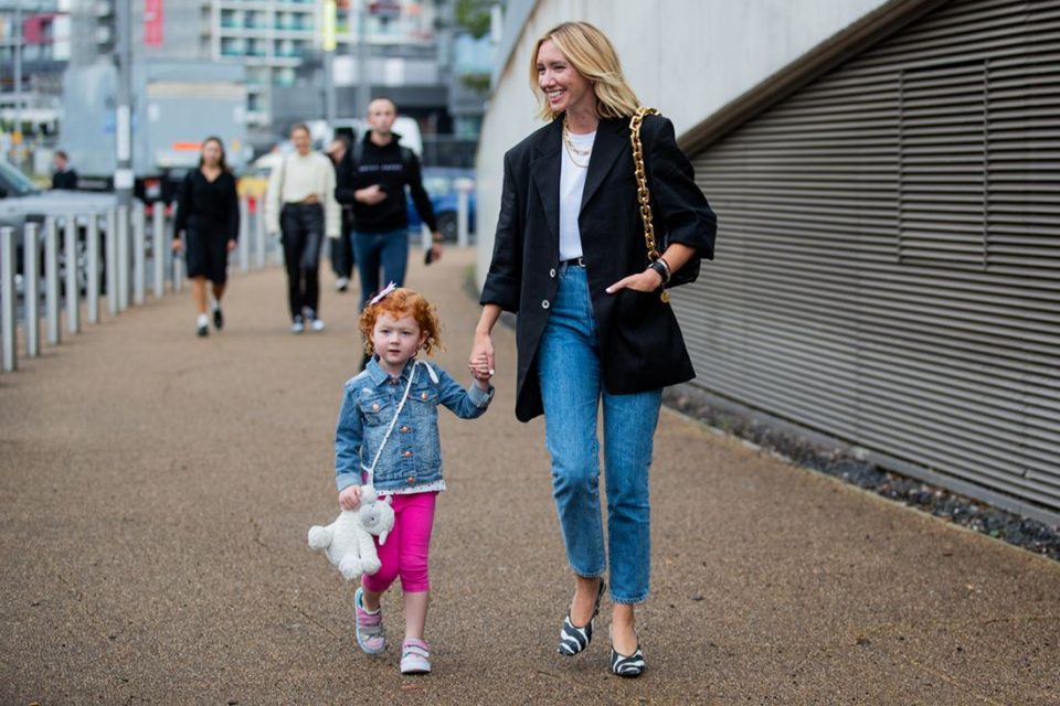 Mulher branca e loira, de cabelo liso longo, andando nas ruas de Londres durante a Semana de Moda. Ela está com a filha, uma menina pequena de no máximo 4 anos, de pele clara e cabelos ruivos cacheados.