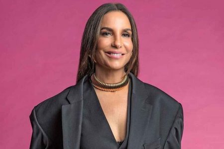 foto colorida de ivete sangalo com blusa e blazer preto sorrindo em fundo rosa - metrópoles