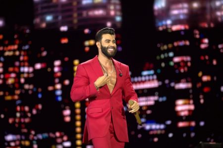 A foto colorida mostra o cantor Gusttavo Lima vestido de terno vermelho segurando um microfone dourado na mão. Ele está em um palco, em frente a um telão que imita luzes de uma cidade