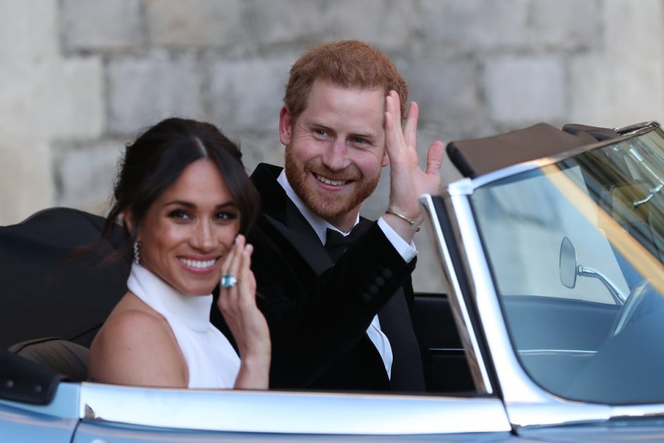 Príncipe Harry e a duquesa Meghan Markle acenam de dentro de um carro conversível, sorrindo - Metrópoles