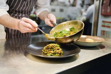 Cozinheiro passa comida de uma panela de metal para prato liso e preto. Ele está em uma cozinha de restaurante - Metrópoles