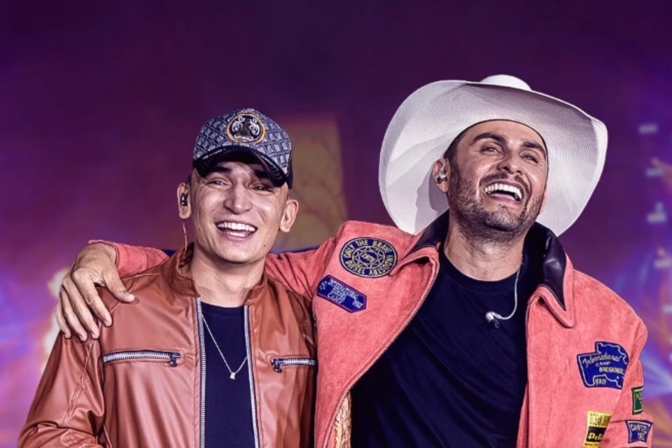 Mano Walter e João Gomes sorriem abraçados em foto. Eles usam jaquetas em tons terrosos e blusas pretas - Metrópoles