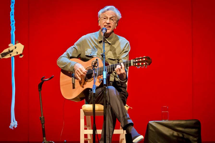 Foto colorida de Caetano Veloso sentado segurando violão - Metrópoles