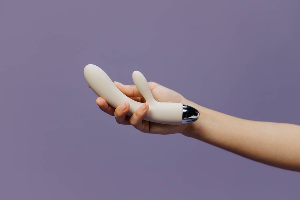 Foto colorida de uma mão feminina segurando um vibrador branco em um fudno roxo - Metrópoles