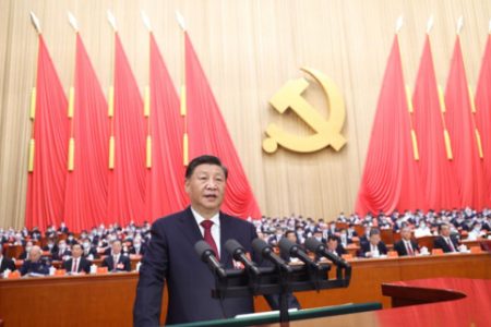 Presidente Xi Jinping da China discursa em sede do Partido Comunista Chinês, de costas para a plateia. Ao fundo, bandeiras vermelhas e o símbolo do comunismo, uma foice com martelo - Metrópoles