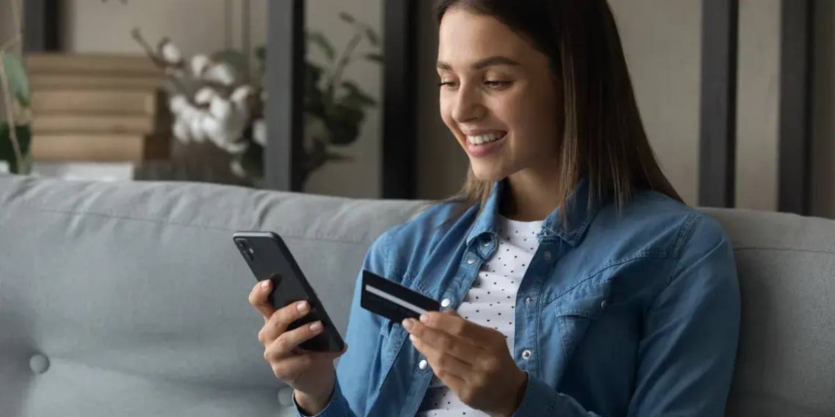 Mulher sorrindo e sentada em sofá segura cartão de crédito e celular nas mãos - Metrópoles