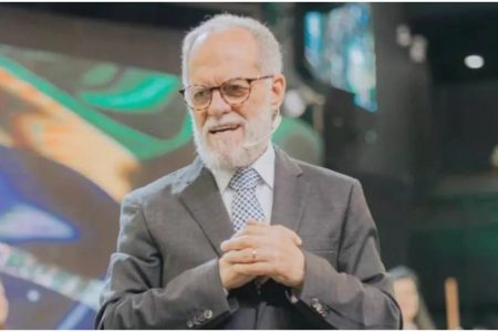 Márcio Valadão, pai do também pastor André Valadão durante culto. Ele usa terno, óculos e microfone e fala para uma plateia com bandeira do Brasil em telão atrás - Metrópoles