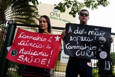 mulher e homem seguram cartazes em frente ao predío do conselho federal de medicina. Nos cartazes há as frases "cannabis é ciência e medicina e saúde" e "a dor não espera"