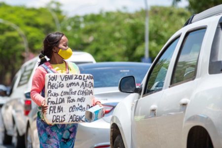 Mulher venezuelana com vestido colorido e máscara segurando placa com os dizeres: "Sou venezuelana. Preciso uma ajuda para comprar comida, fralda para minha filha. Obrigado. Deus abençoe", circula entre carros
