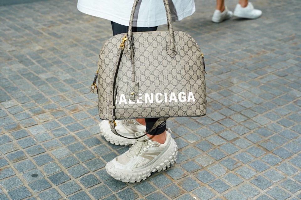 Pessoa andando pelas ruas de Paris com tênis branco, de proporção grande, da marca Balenciaga. Usa ainda uma calça legging preta, uma camiseta branca longa e uma bolsa de couro cinza também da Balenciaga.