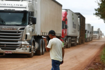 Caminhoneiro ouve aúdio em celular enquanto caminha por beira de rodovia onde se vê vários caminhos parados por conta de bloqueios realizados por apoiadores do presidente derrotado nas eleições, Jair Bolsonaro - Metrópoles