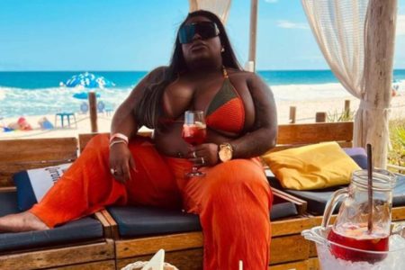 Jojo Todynho vestida de biquini laranja e calça laranja de oculos escuros segurando taça sentada em banco de madeira na praia - Metrópoles