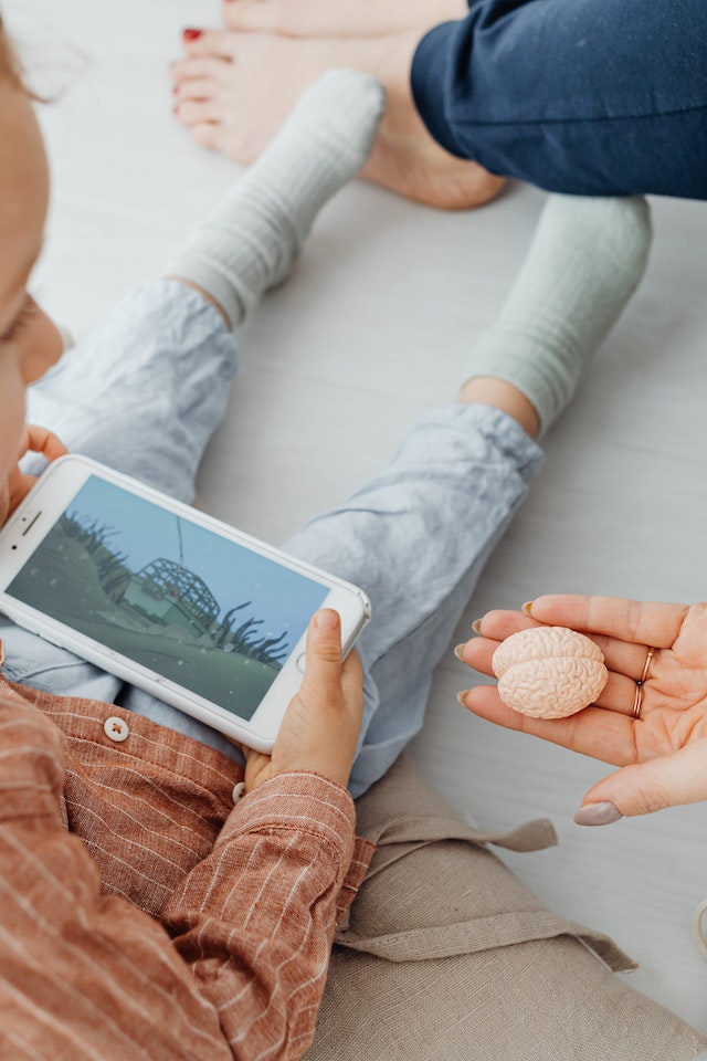 Criança com tablet nas mãos e uma mão adulta segurando um cérebro de plástico ao lado