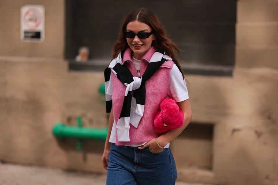 Mulher jovem e branca, de cabelo castanho ondulado, andando pelas ruas de Nova York durante a New Yorker Fashion Week, em 13 de setembro de 2022. Ela usa óculos de sol pretos, camiseta branca, colete de suéter de lã rosa, moletom listrado preto/branco. Uma calça jeans azul e uma mochila fofa rosa Bottega Veneta.