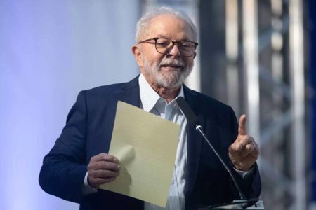 Em imagem colorida, Lula fala ao microfone com uma pasta na mão - Metrópoles