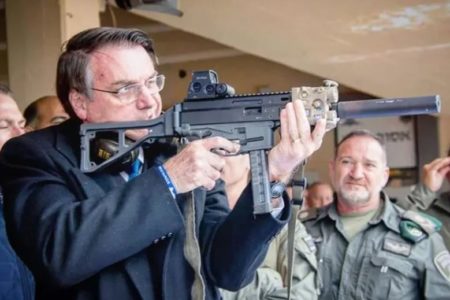 O presidente Bolsonaro mira com fuzil na mão em estande de tira, cercado de homens com roupas militares - Metrópoles