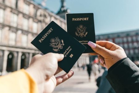 Foto mostra duas mãos segurando um passaporte cada uma, ao fundo mostra uma cidade famosa por seu turismo