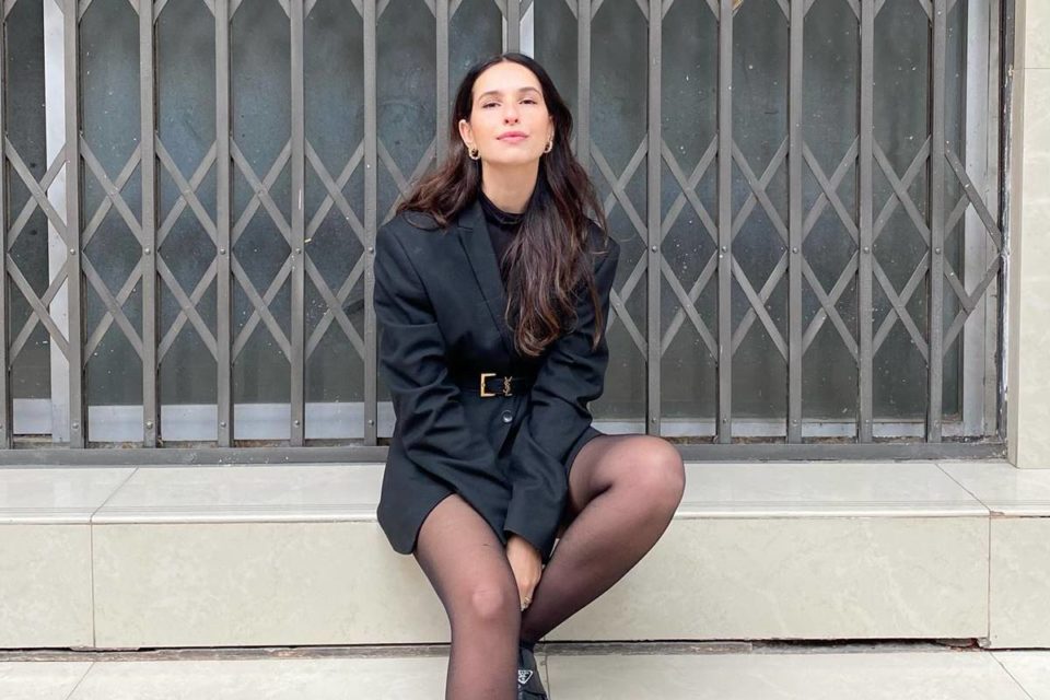 Vic Hollo usa blazer preto com meia calça preta encostada na parede - Metrópoles