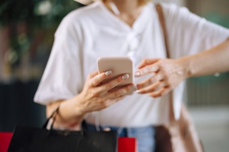 Mulher branca e jovem, de blusa branca, mexendo no celular. Ela segura sacolas de compra preta e vermelho