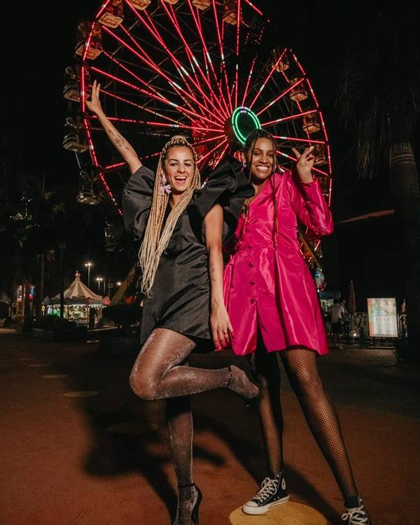 Modelos usam peças da marca 1Quatro. Uma delas usa vestido preto com tecido brilhoso. Outra usa vestido rosa com tecido brilhoso e drapeado - Metrópoles