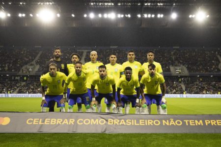 Jogadores da seleção do Brasil em pose de formação para foto em campo, com arquibancada de estádio ao fundo. Eles usam camisa amarela e short azul - Metrópoles