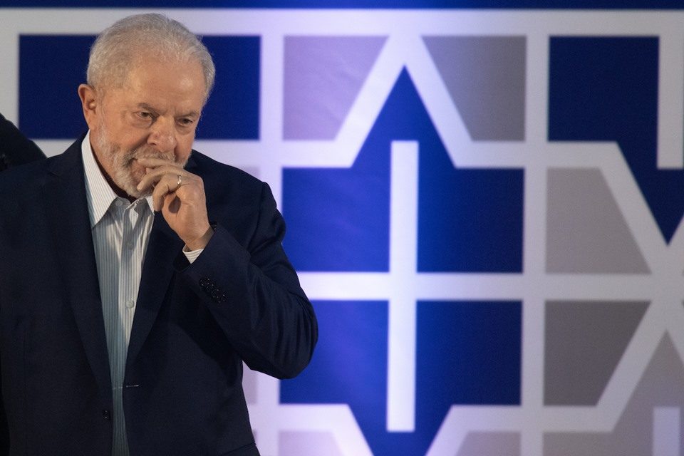 Imagem colorida mostra o presidente eleito Lula diante de um mural multicolorido. Ele está com uma das mãos na boca, dando a impressão de estar em reflexão - Metrópoles