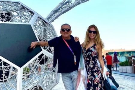 Galvão Bueno e a esposa Desirée. Ele está com uma camisa preta e calça jeans, ela está com um vestido preto florido. Os dois estão com óculos escuros e posam ao lado de uma escultura de bola de futebol - Metrópoles