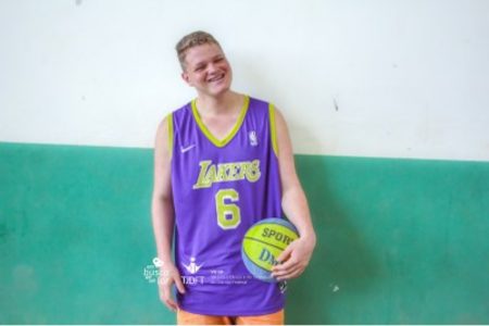 Foto de um menino loiro com blusa regato roxa, segurando uma bola