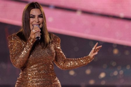 A foto colorida mostra a cantora Naiara Azevedo cantando segurando um microfone. Ela usa um vestido de mangas compridas feito de paetê dourado