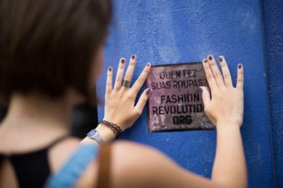 Na imagem com cor, mulher branca cola cartaz em parede azul - Metrópoles