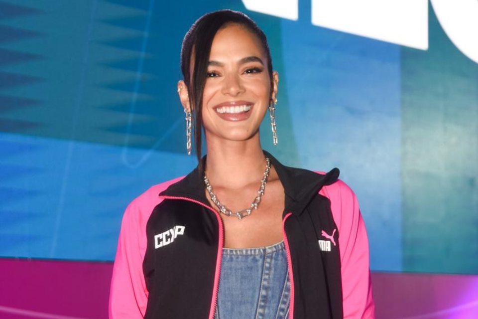 Foto colorida de Bruna Marquezine. Ela usa brincos extravagantes em cor prata, um colar prata, um casaco da CCXP preto com rosa, uma blusa jeans e está sorrindo - Metrópoles