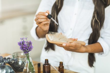 Mulher negra segura uma escova de cabelo com uma das mãos e, na oura, segura o conta-gotas de óleo natural. Parece a preparação de um tratamento capilar. - Metrópoles