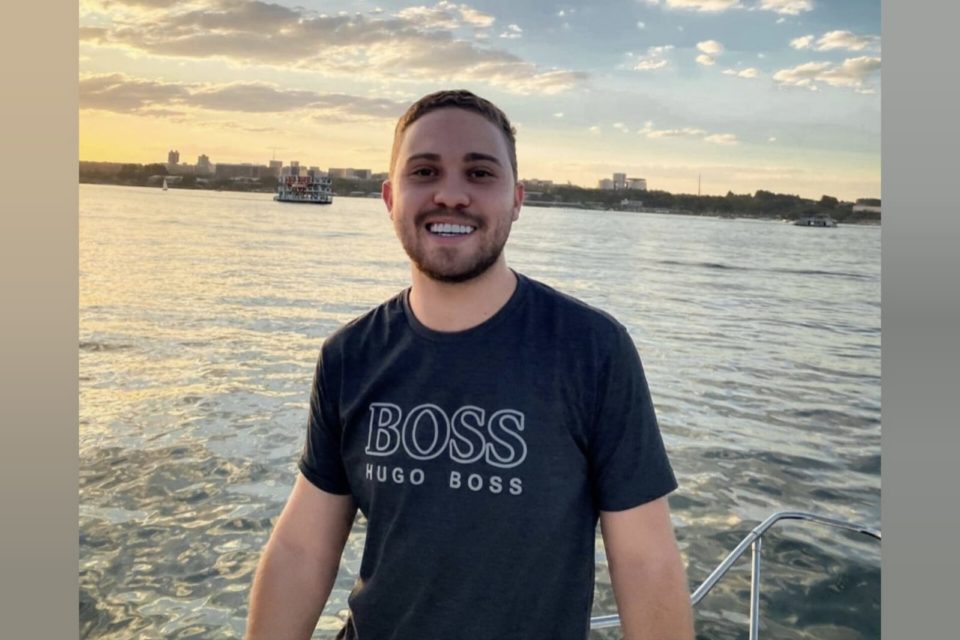 Imagem colorida de homem com camisa preto, com um lago no fundo