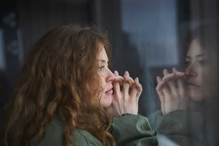 Mulher branca de cabelos ruivos e cacheados está olhando pensativa pela janela, que reflete o seu olhar triste. A imagem representa uma pessoa com depressão - Metrópoles
