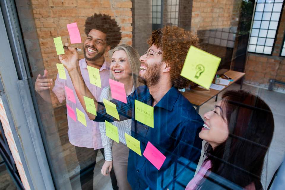 Na foto, há quatro pessoas sorrindo em frente a um vidro com post-its