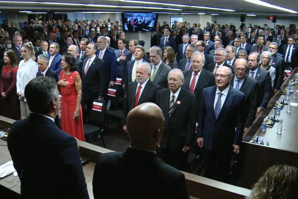 O presidente e vice eleitos Lula e Alckmin em meio a outros políticos e membros do governo de transição na posse do ministro do TCU, Bruno Dantas. Eles aparecem de pé na plateia de frente pra mesa com os ministros - Metrópoles