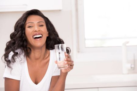 Mulher vestindo branco posando com copo d'água na mão e sorrindo
