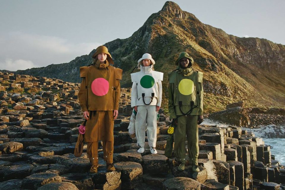 Três homens posando para foto em uma praia com várias pedras e um morro ao fundo. Um é branco europeu, outro é asiático e o terceiro é negro. Eles usam roupas para prática esportiva no frio da marca Moncler. - Metrópoles