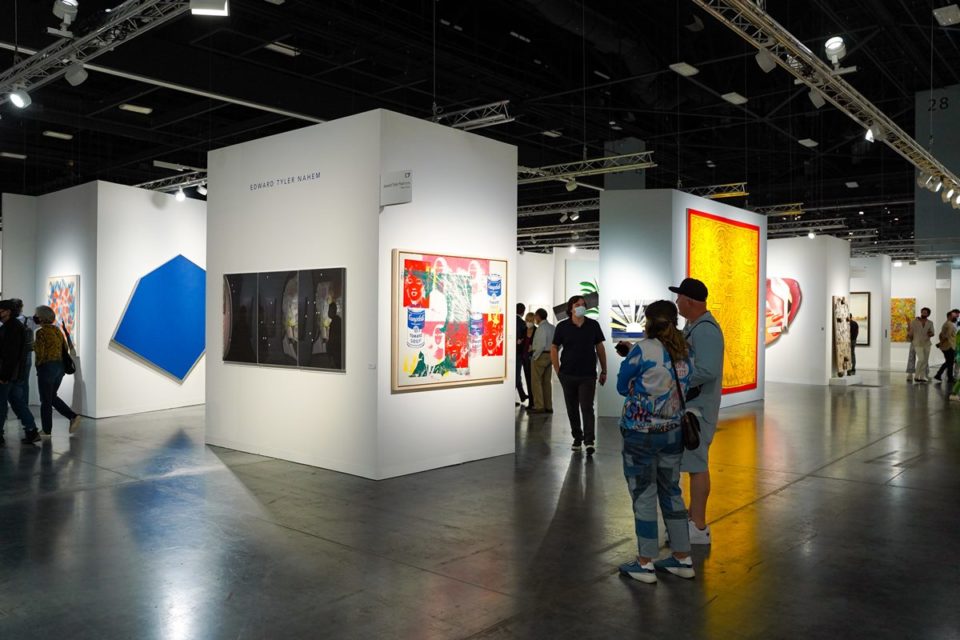 Um homem e uma mulher, ambos brancos e de meia idade, admiram uma obra em uma parede de uma exposição de arte. A foto é do evento Art Basel, em Miami, nos EUA, em 2021. Ela usa uma jaqueta e uma calça, ambos jeans, um boné e tênis. O homem usa um conjunto de shorts e casaco de moletom e tênis branco. - Metrópoles