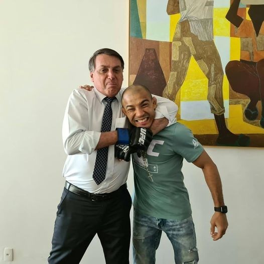 O lutador José Aldo e o presidente Jair Bolsonaro posam para foto sorrindo, com o esportivo recebendo um "golpe" de brincadeira do mandatário - Metrópoles