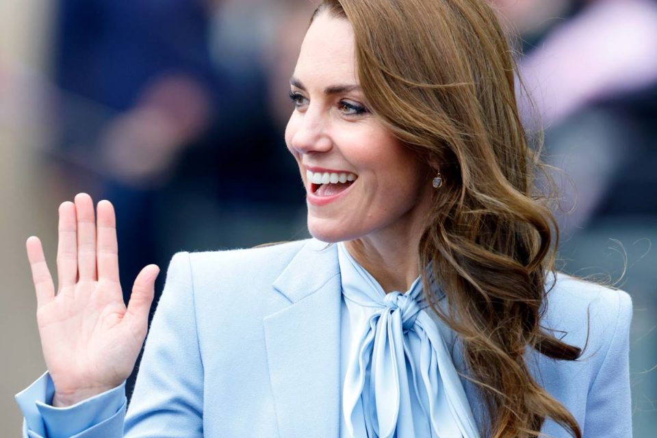 Mulher branca sorrindo em foto, com a mão direita levantada. Ela está sorrindo e usando roupas azul - Metrópoes