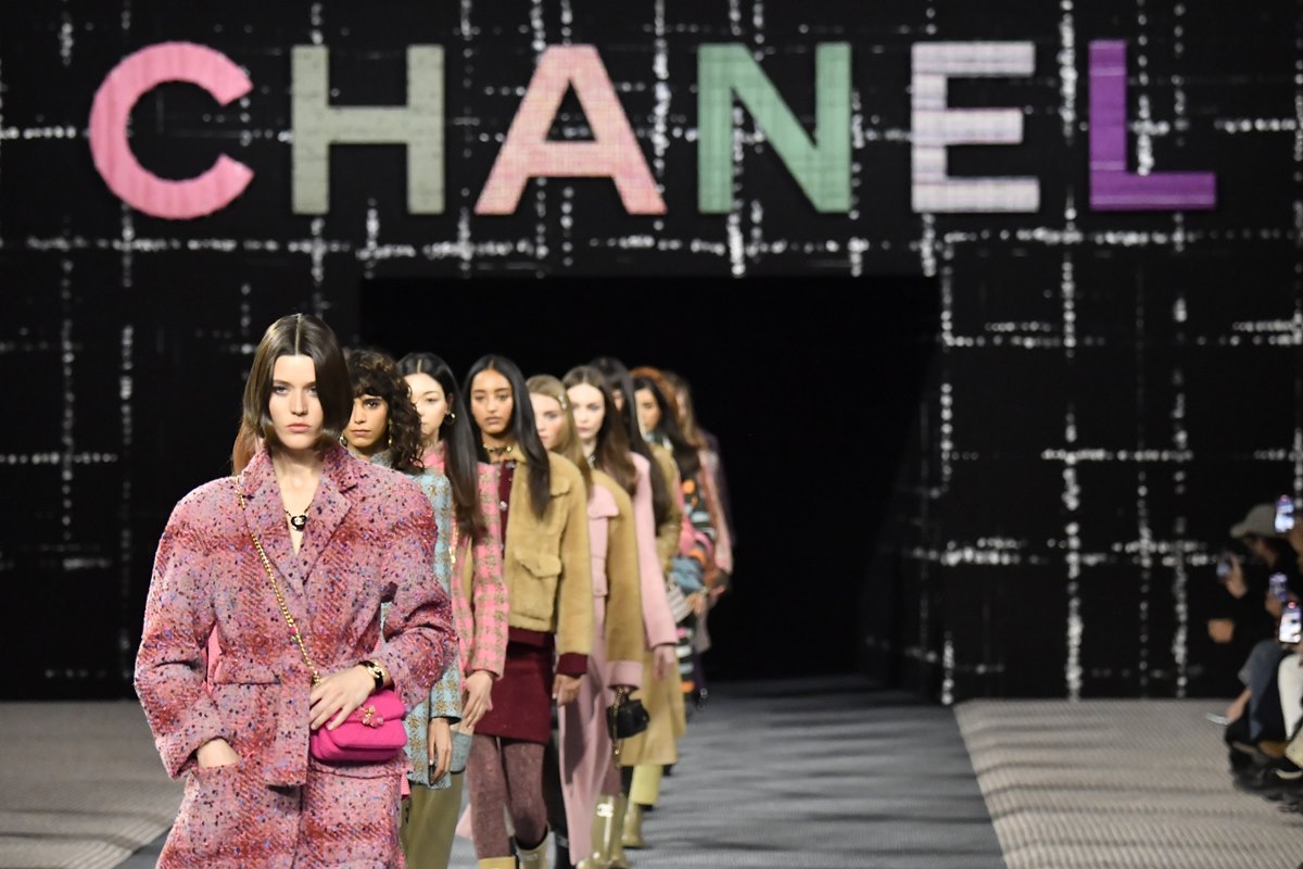 luxo Fila de modelos mulheres e jovem no final do desfile da marca francesa Chanel. Ela usam roupas da marca. No fundo, é possível ver um painel de tecido quadriculado com a logo da marca. - Metrópoles