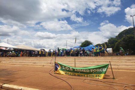 Manifestantes bolsonaristas que resistem no acampamento do QG do Exército aparecem diante de escadaria sentados de baixo de tendas e segurando faixas - Metrópoles