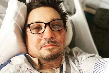 Jeremy Renner em uma cama de hospital. Ele usa óculos de grau e está com o rosto machucado - Metrópoles