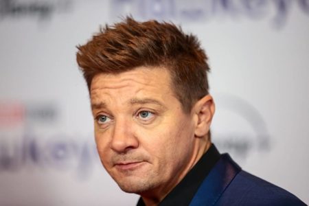 Ator Jeremy Renner vestido de azul, semblante sério no fundo branco com patrocinadores