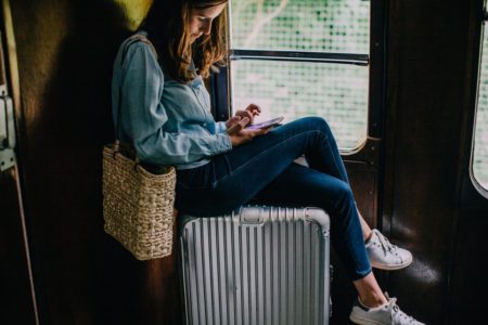 Mulher branca e jovem, de cabelo liso castanho, mexendo no celular dentro de um vagão de trem. Ela usa uma camisa jeans, uma calça jeans, um tênis cinza da Adidas e uma bolsa de palha. Está sentada em cima de uma mala prateada. - Metrópoles