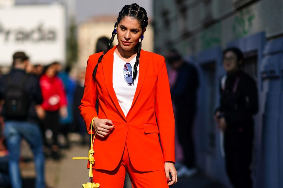 Mulher branca e jovem, de cabelo liso preto amarrado em tranças, posa para foto nas ruas de Milão, na Itália, durante a Semana de Moda. Usa uma camiseta branca e um conjunto de calça e blazer, ambos laranja. - Metrópoles